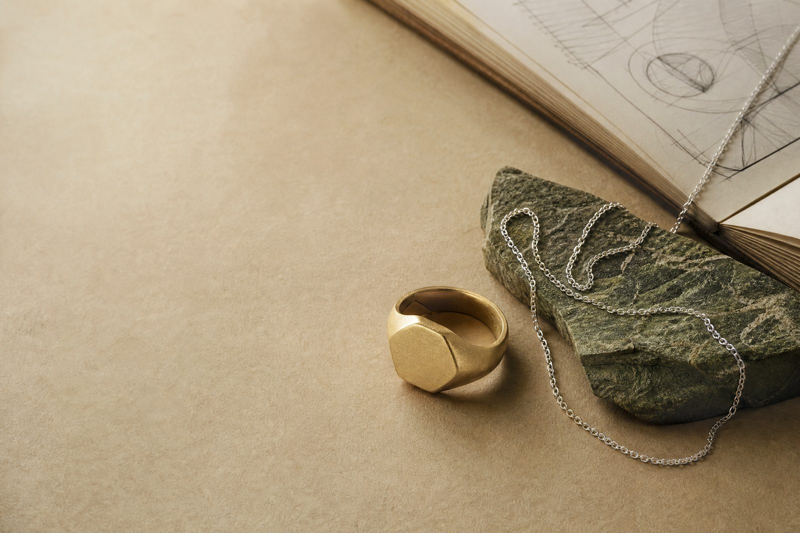 A sculptural signet ring and fine chain arranged on parchment beside raw olive-toned stone
